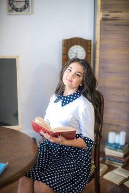 A cute teenage girl with long hair in a classic school dress in a classic interior with books and notebooks at the lesson. School life. Back to school