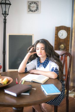 A cute teenage girl with long hair in a classic school dress in a classic interior with books and notebooks at the lesson. School life. Back to school