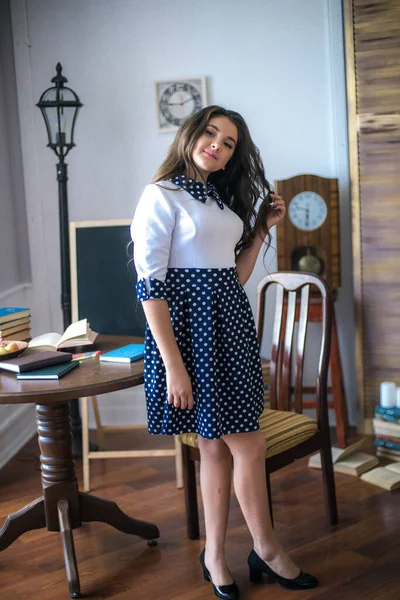 A cute teenage girl with long hair in a classic school dress in a classic interior with books and notebooks at the lesson. School life. Back to school