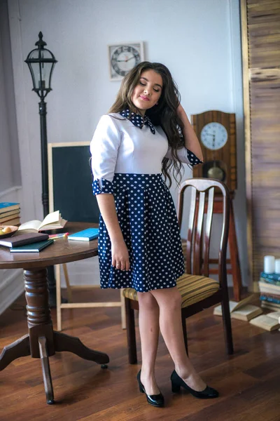 A cute teenage girl with long hair in a classic school dress in a classic interior with books and notebooks at the lesson. School life. Back to school