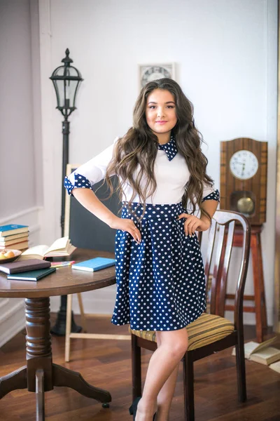 A cute teenage girl with long hair in a classic school dress in a classic interior with books and notebooks at the lesson. School life. Back to school