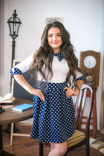 A cute teenage girl with long hair in a classic school dress in a classic interior with books and notebooks at the lesson. School life. Back to school