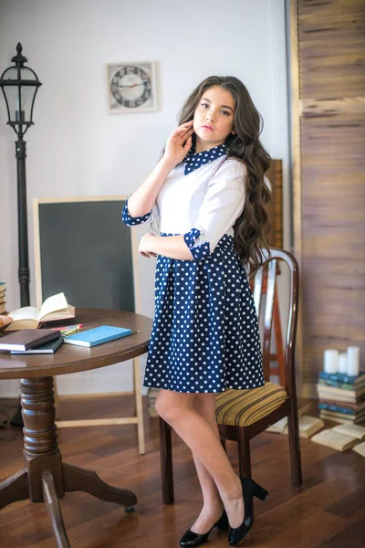 A cute teenage girl with long hair in a classic school dress in a classic interior with books and notebooks at the lesson. School life. Back to school