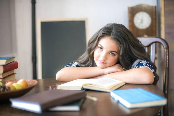 A cute teenage girl with long hair in a classic school dress in a classic interior with books and notebooks at the lesson. School life. Back to school