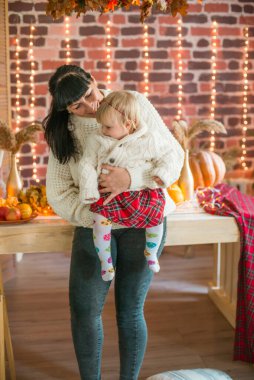 Beyaz örgü süveterler içinde, battaniye, yastık, balkabağı, sonbahar yaprakları ve elmalarla süslenmiş genç bir anne ve kız. Sonbahar havası. Cadılar Bayramı