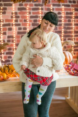 Beyaz örgü süveterler içinde, battaniye, yastık, balkabağı, sonbahar yaprakları ve elmalarla süslenmiş genç bir anne ve kız. Sonbahar havası. Cadılar Bayramı