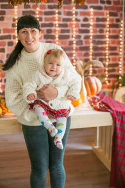 Beyaz örgü süveterler içinde, battaniye, yastık, balkabağı, sonbahar yaprakları ve elmalarla süslenmiş genç bir anne ve kız. Sonbahar havası. Cadılar Bayramı