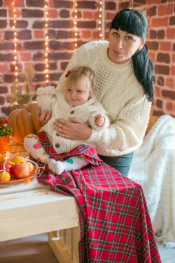 Beyaz örgü süveterler içinde, battaniye, yastık, balkabağı, sonbahar yaprakları ve elmalarla süslenmiş genç bir anne ve kız. Sonbahar havası. Cadılar Bayramı