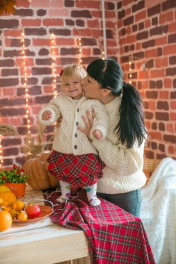 Beyaz örgü süveterler içinde, battaniye, yastık, balkabağı, sonbahar yaprakları ve elmalarla süslenmiş genç bir anne ve kız. Sonbahar havası. Cadılar Bayramı