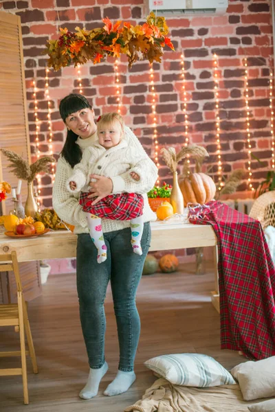 Beyaz örgü süveterler içinde, battaniye, yastık, balkabağı, sonbahar yaprakları ve elmalarla süslenmiş genç bir anne ve kız. Sonbahar havası. Cadılar Bayramı