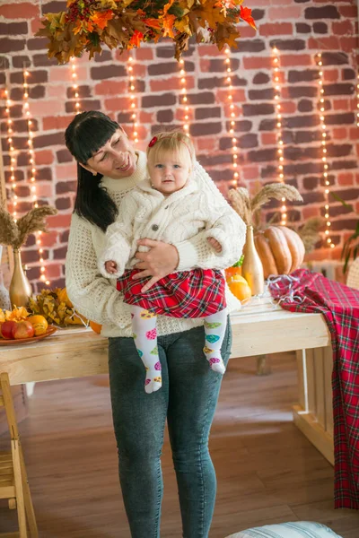 Beyaz örgü süveterler içinde, battaniye, yastık, balkabağı, sonbahar yaprakları ve elmalarla süslenmiş genç bir anne ve kız. Sonbahar havası. Cadılar Bayramı