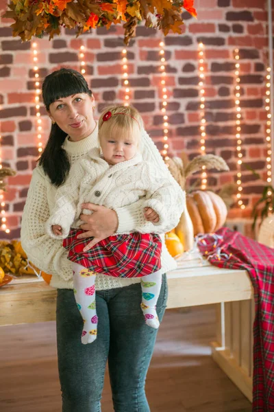 Beyaz örgü süveterler içinde, battaniye, yastık, balkabağı, sonbahar yaprakları ve elmalarla süslenmiş genç bir anne ve kız. Sonbahar havası. Cadılar Bayramı