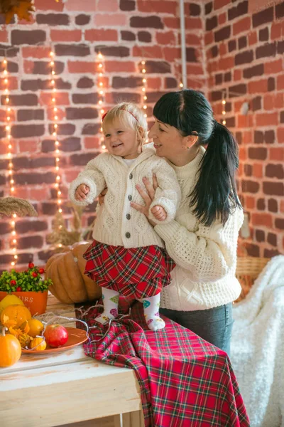 Beyaz örgü süveterler içinde, battaniye, yastık, balkabağı, sonbahar yaprakları ve elmalarla süslenmiş genç bir anne ve kız. Sonbahar havası. Cadılar Bayramı