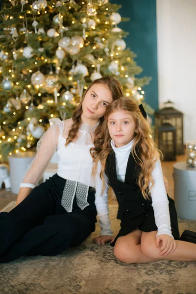 Lovely girls sisters with blonde curly hair with bans to the tols in classic clothes by the Christmas tree  with presents in a classic interior. Christmas mood