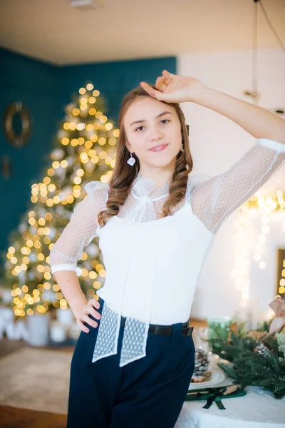 白いブラウスとヘアスタイルとメイクで流行のズボンの魅力的な若い女の子は 休日のテーブルでクリスマスのために装飾された部屋で お祭り気分 クリスマス — ストック写真