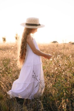 Bir hasır şapka ile beyaz bir elbise ile gün batımında bir yaz alanında sarı uzun saçlı sevimli küçük kız