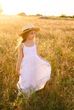 Bir hasır şapka ile beyaz bir elbise ile gün batımında bir yaz alanında sarı uzun saçlı sevimli küçük kız