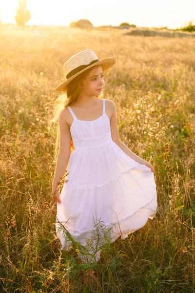 可爱的小女孩与金发长发在夏天的田野在日落与一个白色的礼服与草帽 — 图库照片
