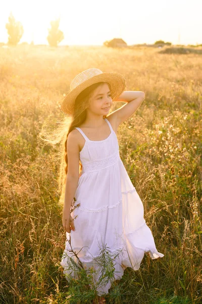 可爱的小女孩与金发长发在夏天的田野在日落与一个白色的礼服与草帽 — 图库照片