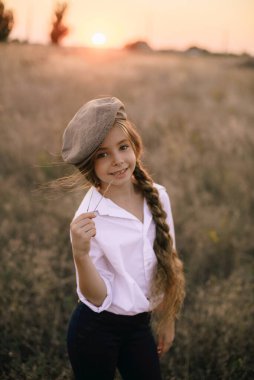 Beyaz gömlekli, uzun sarı saçlı ve yaz tarlasında gün batımında erkek şapkası takan sevimli küçük bir kız..