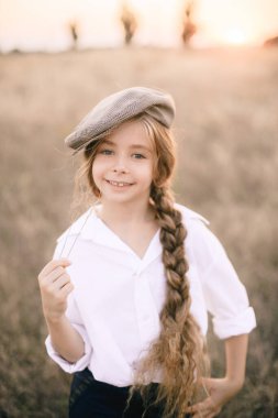 Beyaz gömlekli, uzun sarı saçlı ve yaz tarlasında gün batımında erkek şapkası takan sevimli küçük bir kız..