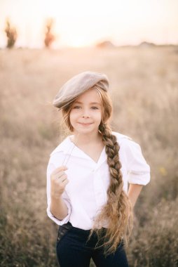 Beyaz gömlekli, uzun sarı saçlı ve yaz tarlasında gün batımında erkek şapkası takan sevimli küçük bir kız..