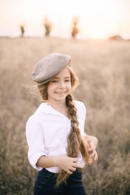Beyaz gömlekli, uzun sarı saçlı ve yaz tarlasında gün batımında erkek şapkası takan sevimli küçük bir kız..
