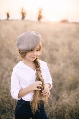 Beyaz gömlekli, uzun sarı saçlı ve yaz tarlasında gün batımında erkek şapkası takan sevimli küçük bir kız..