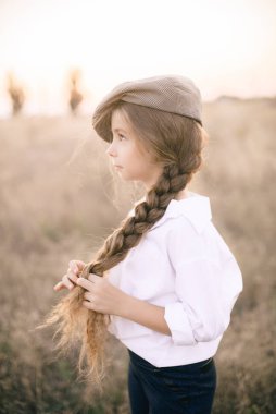 Beyaz gömlekli, uzun sarı saçlı ve yaz tarlasında gün batımında erkek şapkası takan sevimli küçük bir kız..