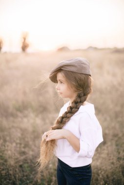 Beyaz gömlekli, uzun sarı saçlı ve yaz tarlasında gün batımında erkek şapkası takan sevimli küçük bir kız..