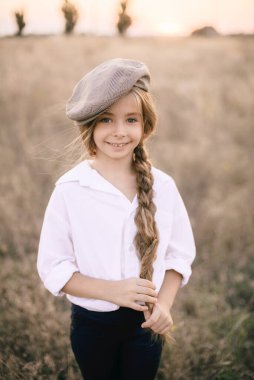 Beyaz gömlekli, uzun sarı saçlı ve yaz tarlasında gün batımında erkek şapkası takan sevimli küçük bir kız..