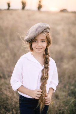 Beyaz gömlekli, uzun sarı saçlı ve yaz tarlasında gün batımında erkek şapkası takan sevimli küçük bir kız..
