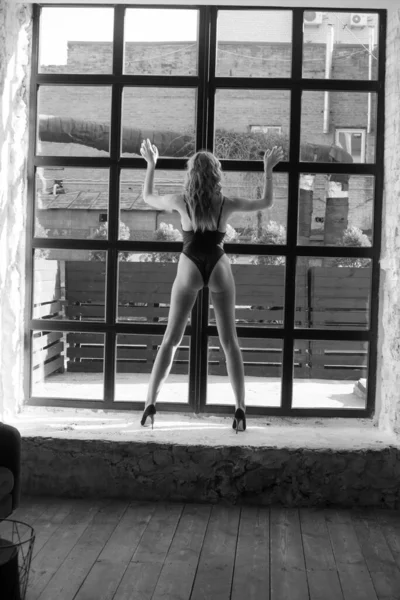 迷人的身材苗条的小女孩 金发卷曲 穿着一件黑色单件泳衣 站在一个大窗户前 美与时尚 — 图库照片