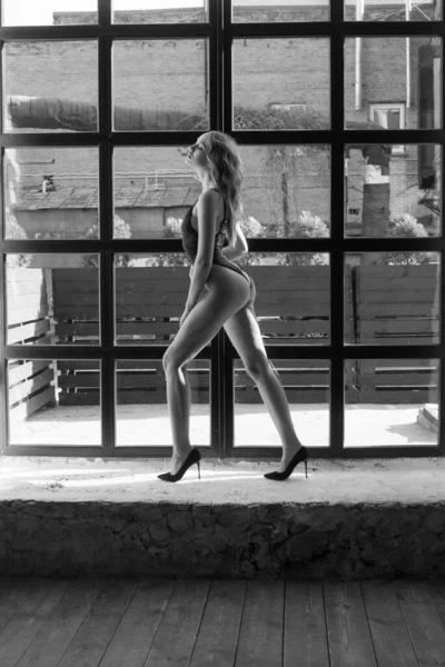 迷人的身材苗条的小女孩 金发卷曲 穿着一件黑色单件泳衣 站在一个大窗户前 美与时尚 — 图库照片