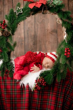 Kırmızı takım elbiseli ve şapkalı şirin bir bebek Noel süslemesinde uyuyor. Noel havası. Mutlu çocukluk.
