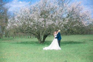 Gelin ve damat sarı uzun saçlı, bahar bahçesinde çiçek açan bir ağacın yanında beyaz elbiseli. Duygular ve duygular