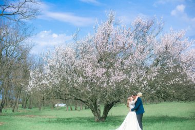 Gelin ve damat sarı uzun saçlı, bahar bahçesinde çiçek açan bir ağacın yanında beyaz elbiseli. Duygular ve duygular