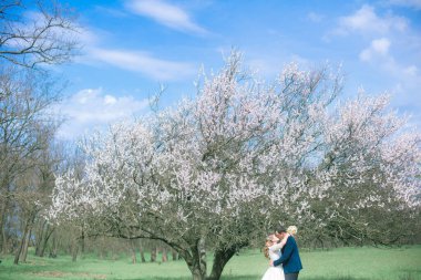 Gelin ve damat sarı uzun saçlı, bahar bahçesinde çiçek açan bir ağacın yanında beyaz elbiseli. Duygular ve duygular