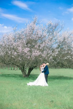 Gelin ve damat sarı uzun saçlı, bahar bahçesinde çiçek açan bir ağacın yanında beyaz elbiseli. Duygular ve duygular
