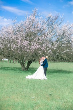 Gelin ve damat sarı uzun saçlı, bahar bahçesinde çiçek açan bir ağacın yanında beyaz elbiseli. Duygular ve duygular