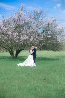 Gelin ve damat sarı uzun saçlı, bahar bahçesinde çiçek açan bir ağacın yanında beyaz elbiseli. Duygular ve duygular