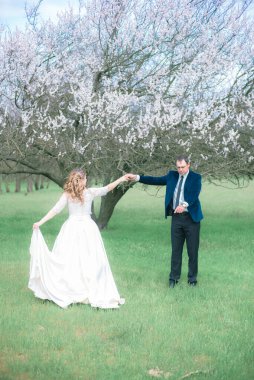 Gelin ve damat sarı uzun saçlı, bahar bahçesinde çiçek açan bir ağacın yanında beyaz elbiseli. Duygular ve duygular