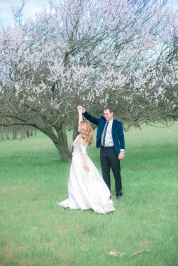 Gelin ve damat sarı uzun saçlı, bahar bahçesinde çiçek açan bir ağacın yanında beyaz elbiseli. Duygular ve duygular