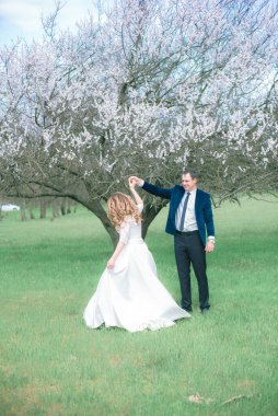 Gelin ve damat sarı uzun saçlı, bahar bahçesinde çiçek açan bir ağacın yanında beyaz elbiseli. Duygular ve duygular