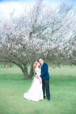 Gelin ve damat sarı uzun saçlı, bahar bahçesinde çiçek açan bir ağacın yanında beyaz elbiseli. Duygular ve duygular