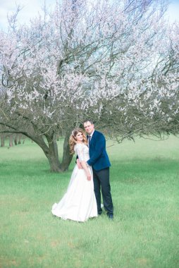 Gelin ve damat sarı uzun saçlı, bahar bahçesinde çiçek açan bir ağacın yanında beyaz elbiseli. Duygular ve duygular