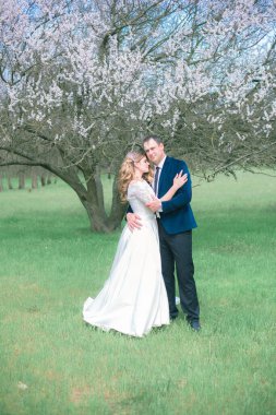 Gelin ve damat sarı uzun saçlı, bahar bahçesinde çiçek açan bir ağacın yanında beyaz elbiseli. Duygular ve duygular