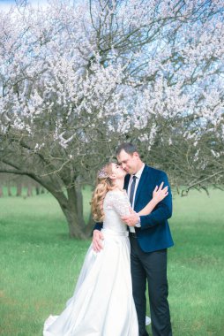 Gelin ve damat sarı uzun saçlı, bahar bahçesinde çiçek açan bir ağacın yanında beyaz elbiseli. Duygular ve duygular