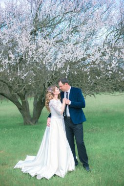 Gelin ve damat sarı uzun saçlı, bahar bahçesinde çiçek açan bir ağacın yanında beyaz elbiseli. Duygular ve duygular