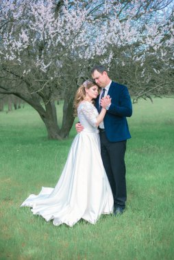 Gelin ve damat sarı uzun saçlı, bahar bahçesinde çiçek açan bir ağacın yanında beyaz elbiseli. Duygular ve duygular